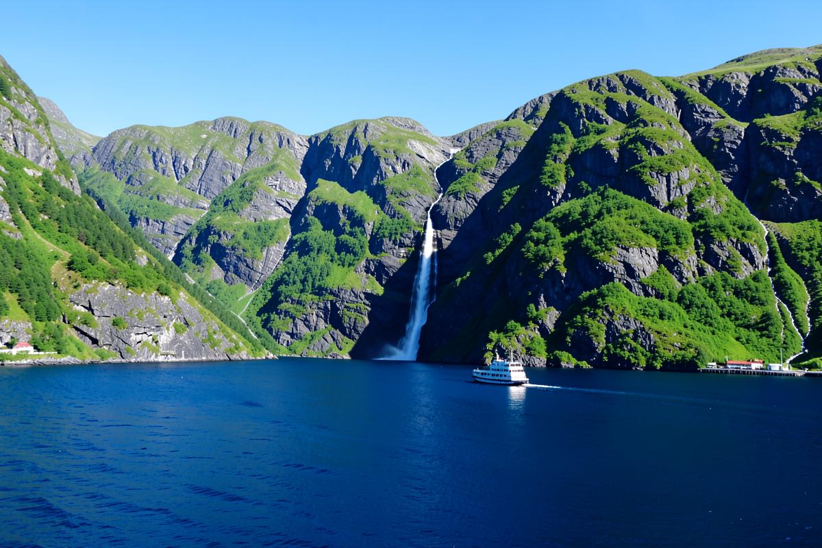 Steile grüne Fjordwände mit Wasserfall und kleinem Schiff auf tiefblauem Wasser