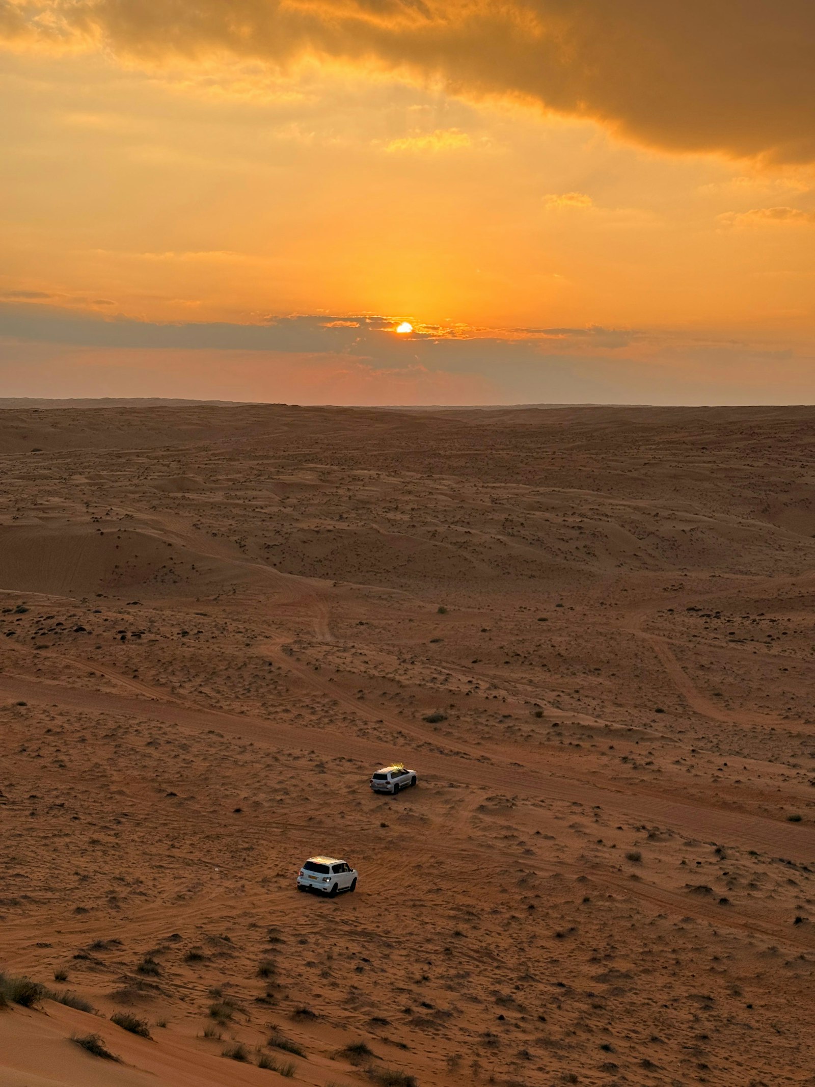 Zwei weiße Geländewagen fahren bei Sonnenuntergang durch eine weite Wüste